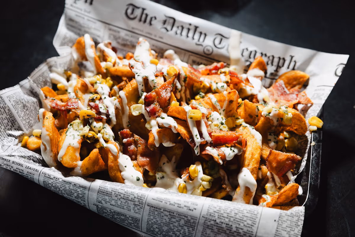 Tray of seasoned potato wedges topped with bacon, corn, herbs, and drizzled with white sauce served on newspaper-style paper.