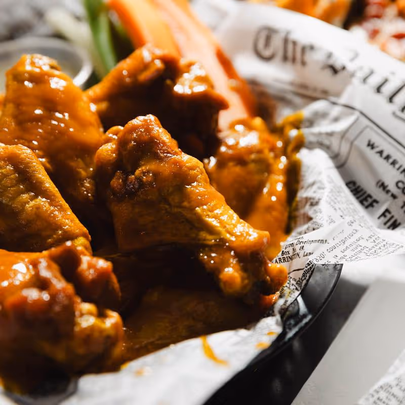 Close-up of a basket filled with glazed chicken wings and some carrot sticks wrapped in newspaper-style paper.