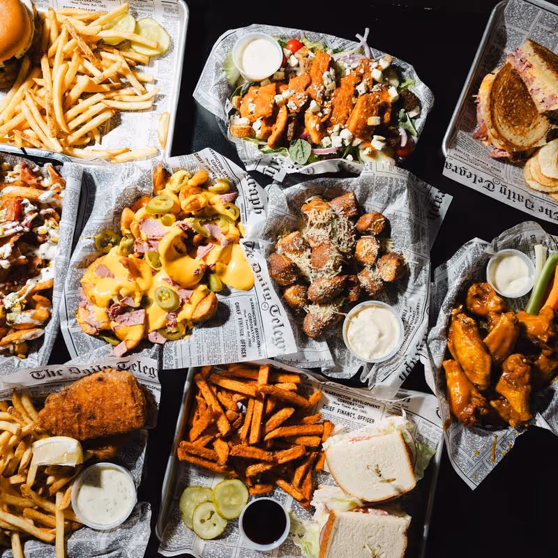 Variety of fast food dishes including cheeseburger with fries, loaded fries with cheese and jalapeños, sweet potato fries, chicken wings, a sandwich, and dipping sauces on trays lined with newspaper-themed paper.