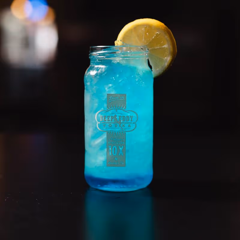 Blue cocktail with ice in a Deep Eddy Vodka mason jar glass garnished with a lemon slice.