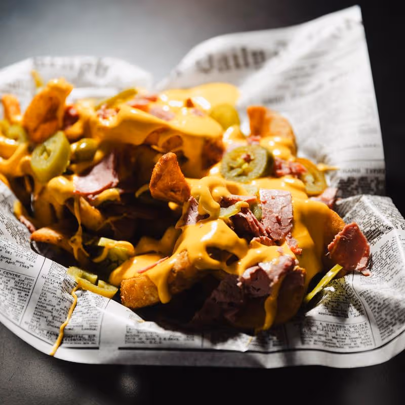 Loaded fries topped with melted cheese, slices of jalapeños, and pieces of corned beef served in a basket lined with newspaper-style wax paper.