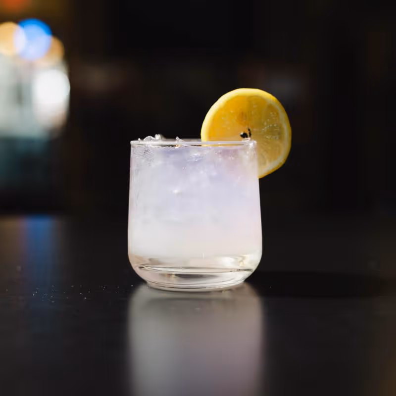 Glass of iced clear drink garnished with a lemon slice on a dark reflective surface.