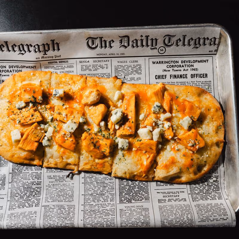 Flatbread pizza topped with chunks of cheese and herbs on a tray lined with a vintage newspaper.