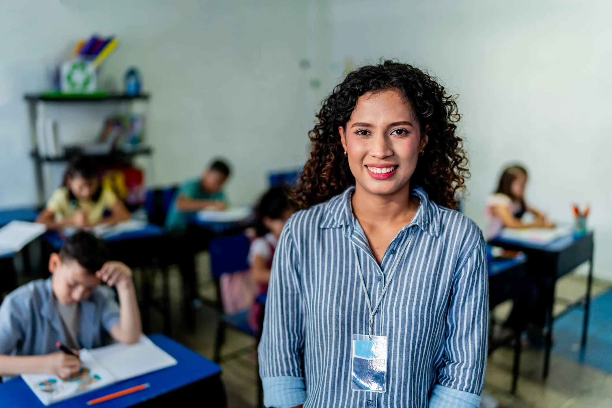 Smiling female teacher with curly hair and striped shirt standing in a classroom with young students working at desks in the background.