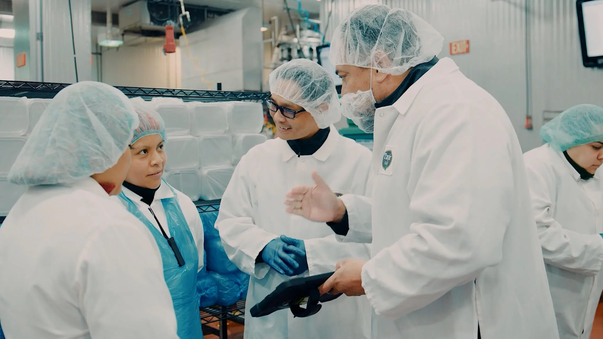 Factory workers wearing protective gear discussing production processes on a tablet inside a food processing facility.