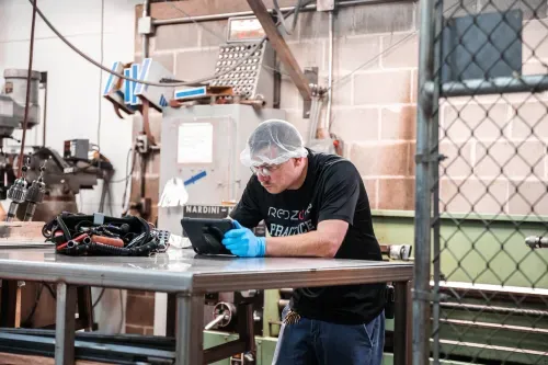 Technician wearing safety gloves and a hairnet reviewing production data on a tablet at a metal workbench.