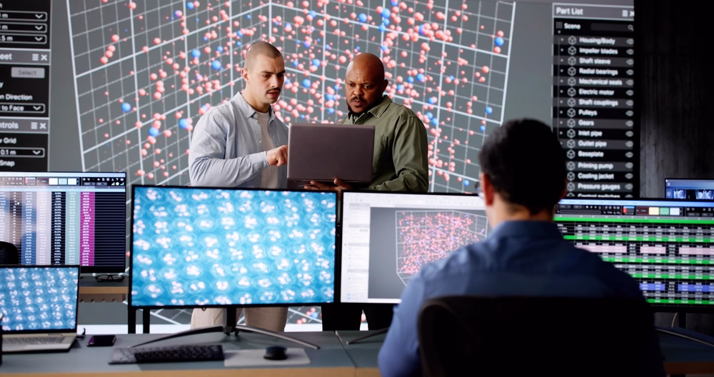 Team of data scientists analyzing complex datasets on multiple screens, with two colleagues reviewing results on a laptop in front of a large visualization of data points.
