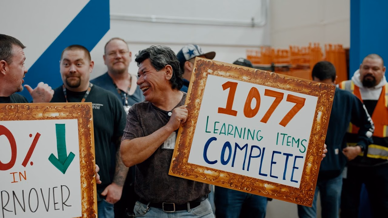 Group of factory workers celebrating achievements, holding framed signs showing completed learning items and performance improvements.