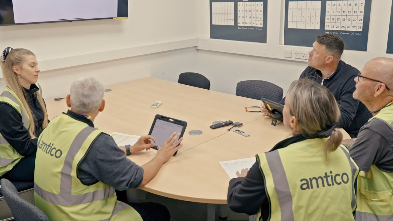 Factory team in high-visibility vests gathered around a table reviewing performance data during a production meeting.