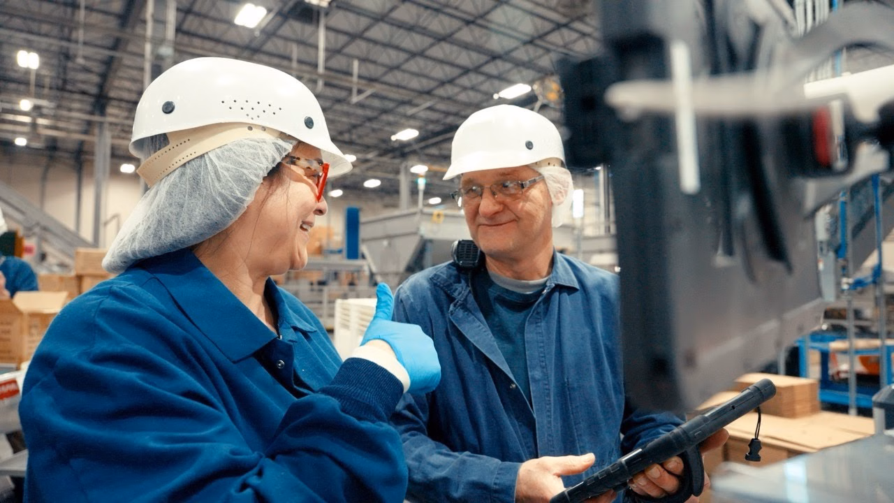 Two factory workers in safety gear smiling and discussing data displayed on a tablet in an industrial setting