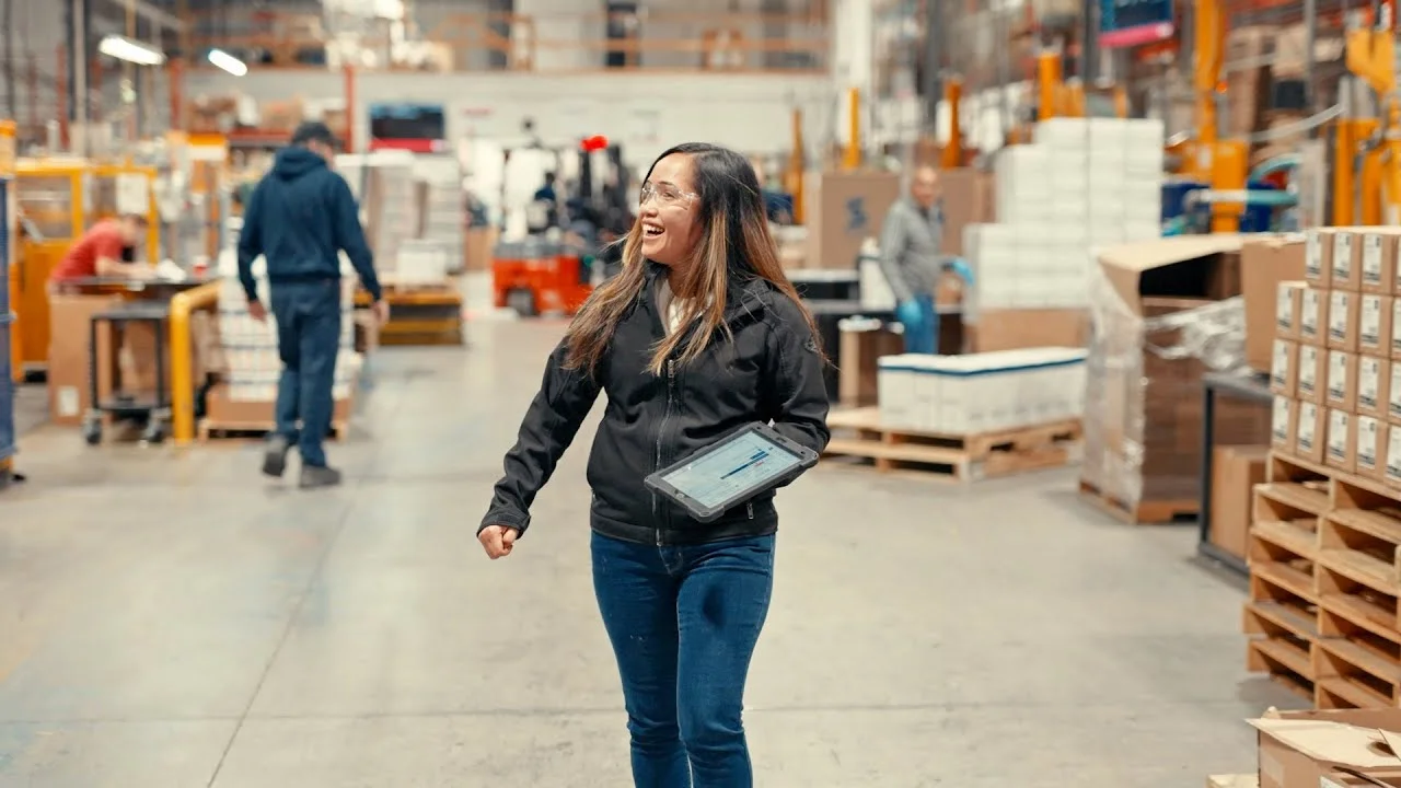 Worker walking through a busy warehouse holding a tablet, surrounded by shelves, pallets, and forklifts.