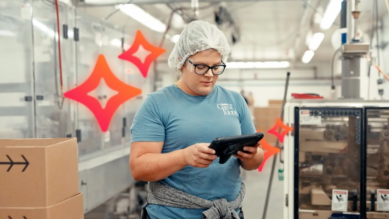 Factory worker wearing a hairnet and glasses using a tablet beside industrial machinery in a production facility.