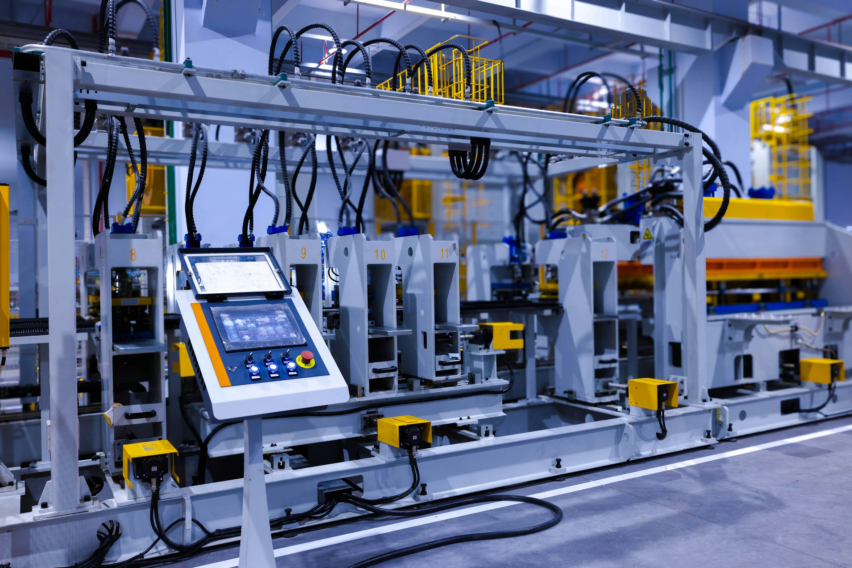 An automated industrial production line featuring a digital control panel interface in the foreground, connected to heavy machinery with numbered stations and hydraulic systems in a modern manufacturing facility.