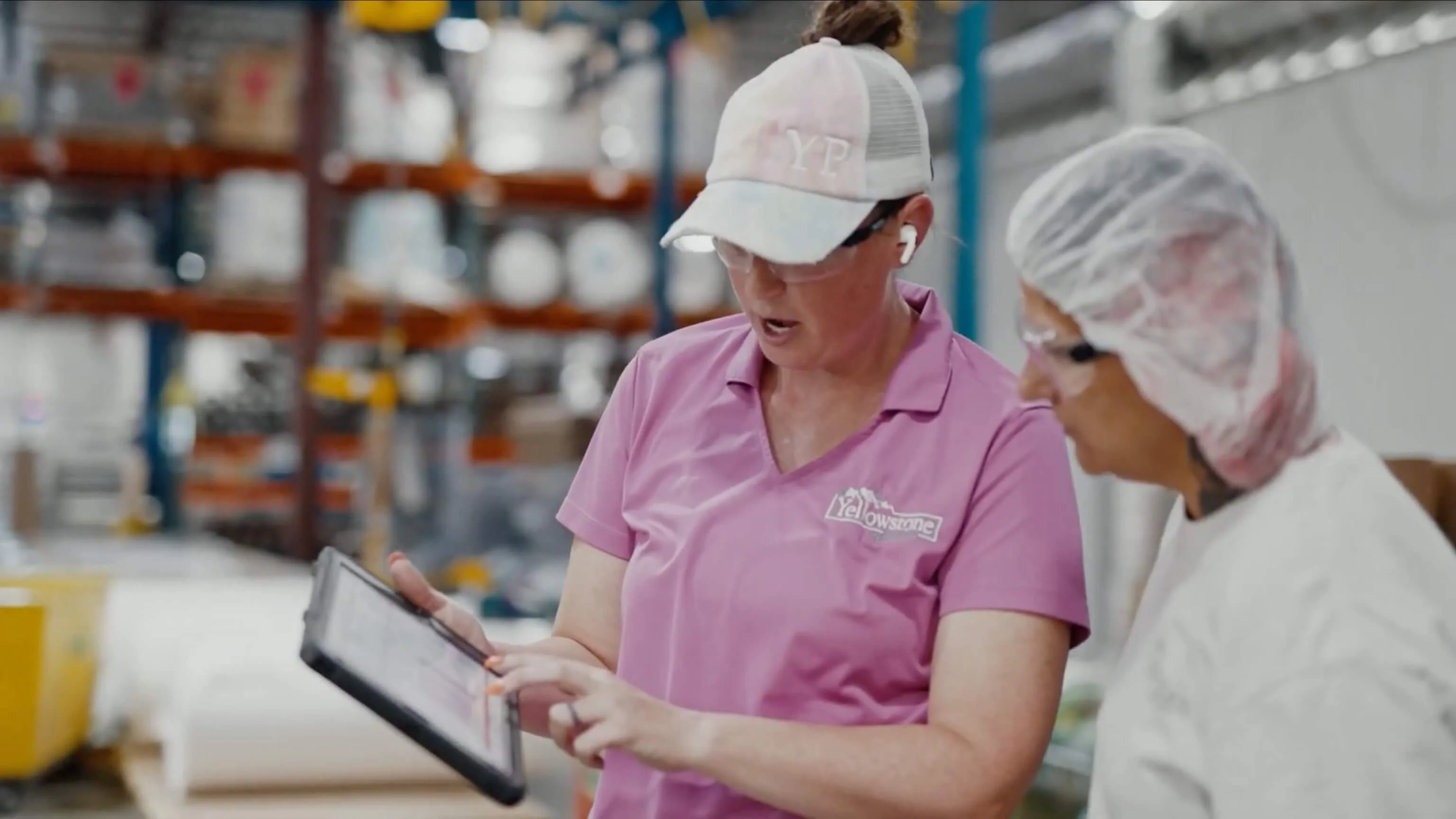 Two factory workers wearing safety gear review information on a tablet in an industrial warehouse setting.