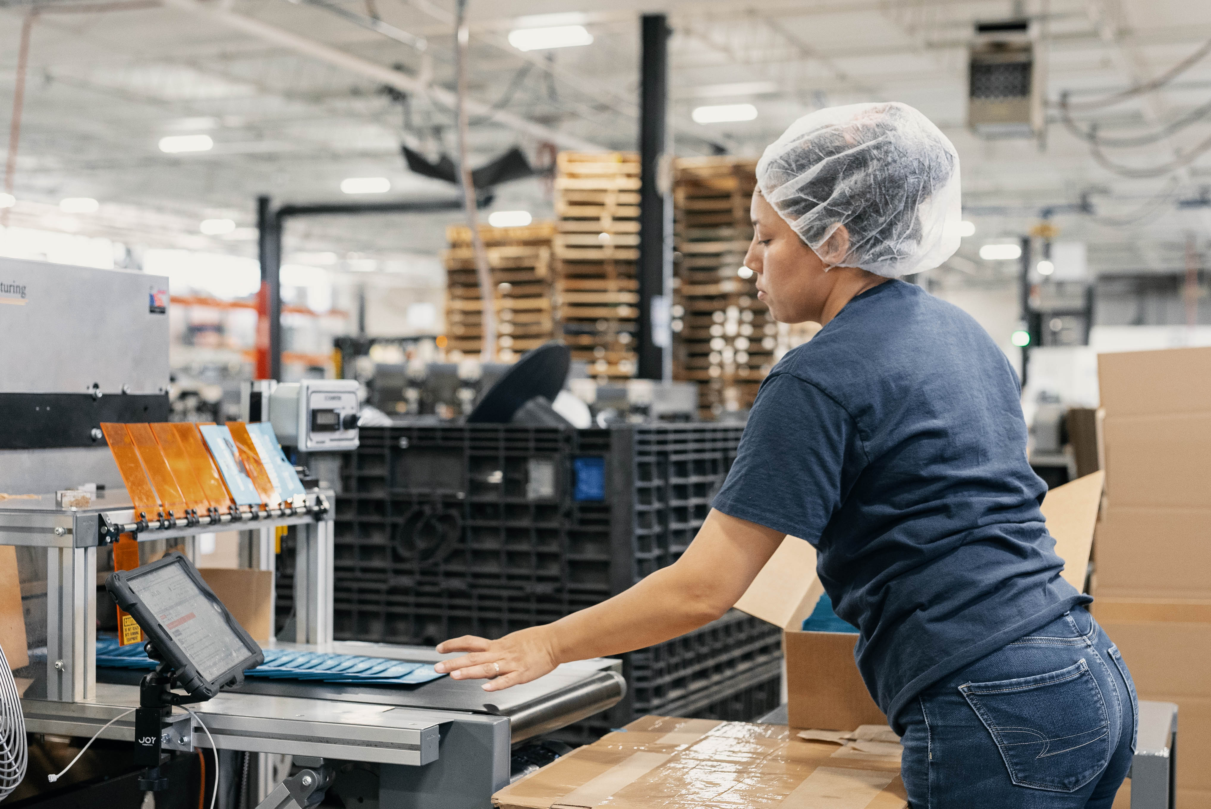 Factory worker wearing hairnet reviews information on a tablet in an industrial warehouse setting.