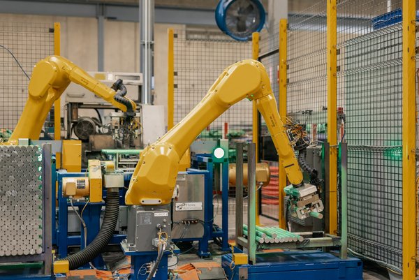 Two bright yellow robotic arms operating inside a secured wire-mesh safety enclosure to sort and handle cylindrical metal components on a factory floor.