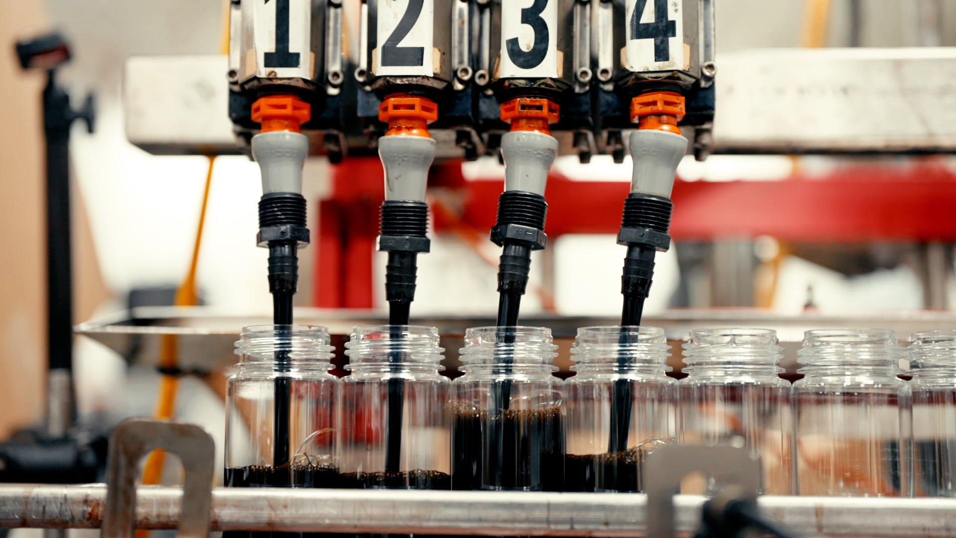 An automated liquid filling machine with four numbered nozzles dispensing a dark liquid into clear plastic bottles on an industrial production line.
