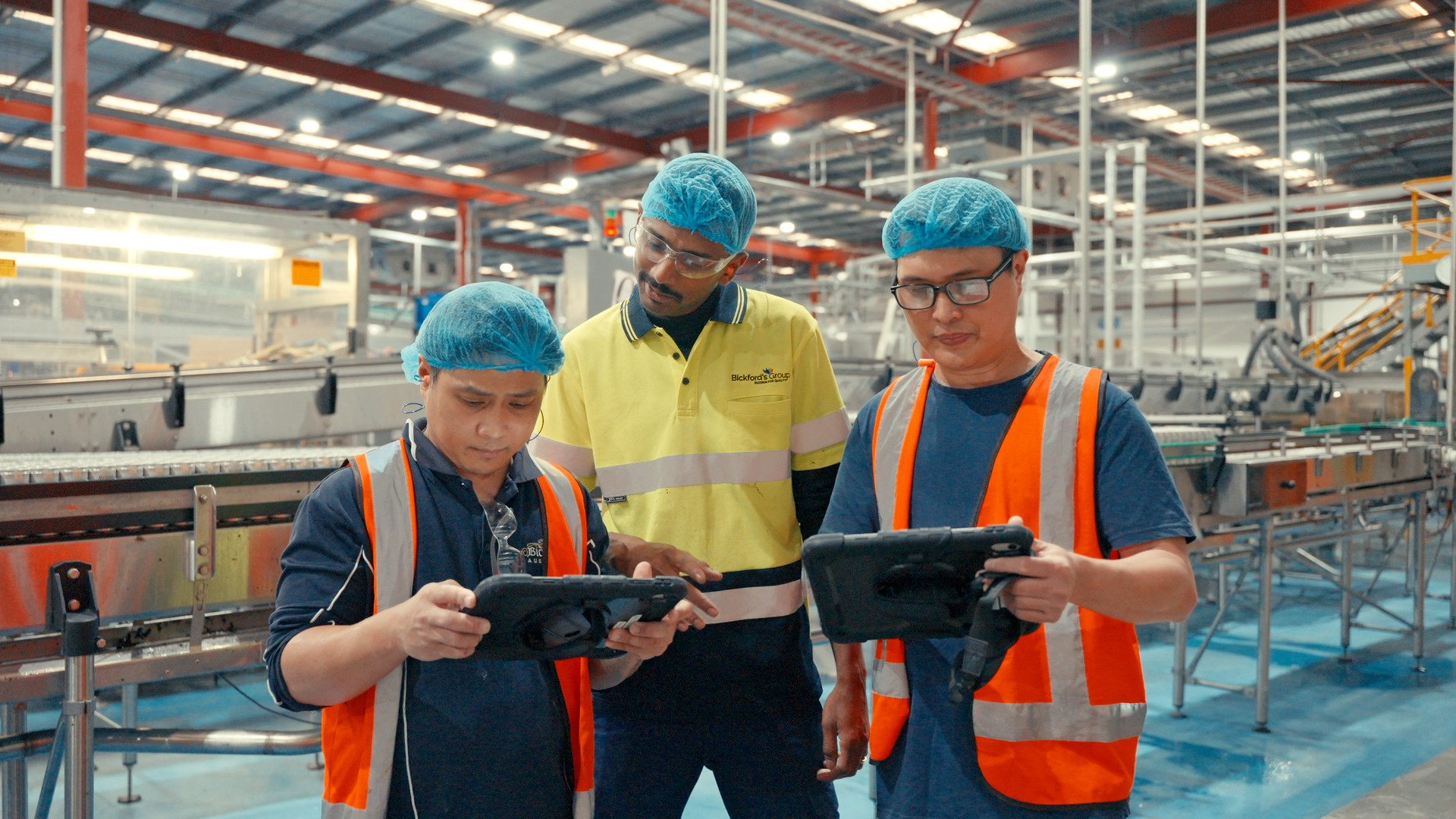 Three manufacturing workers wearing blue hairnets and high-visibility safety vests collaborate on a factory floor while reviewing data on industrial digital tablets.