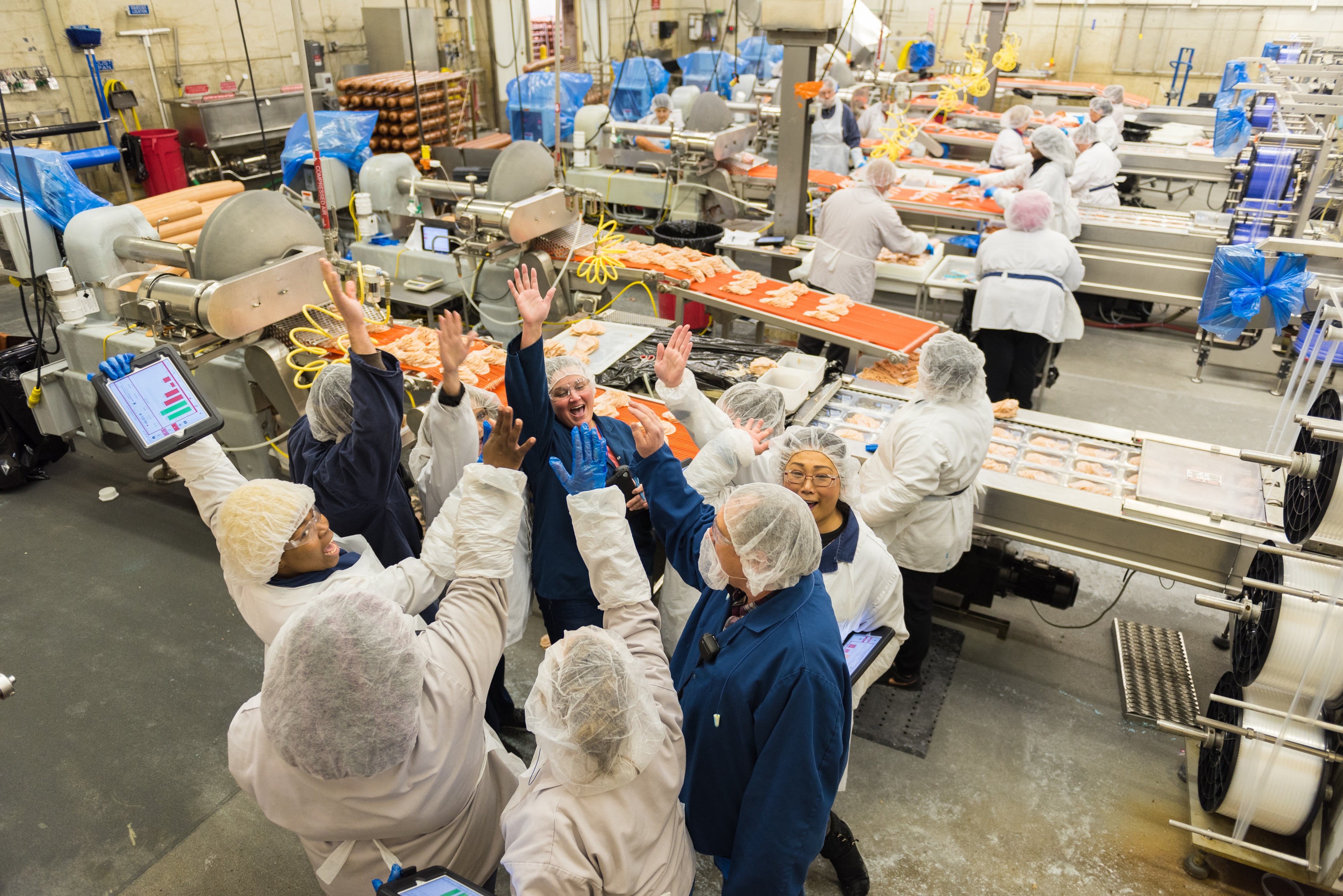 A group of food processing facility workers wearing hairnets and lab coats celebrate in a huddle, giving high-fives and holding tablets with performance charts, on a busy poultry production and packaging floor.
