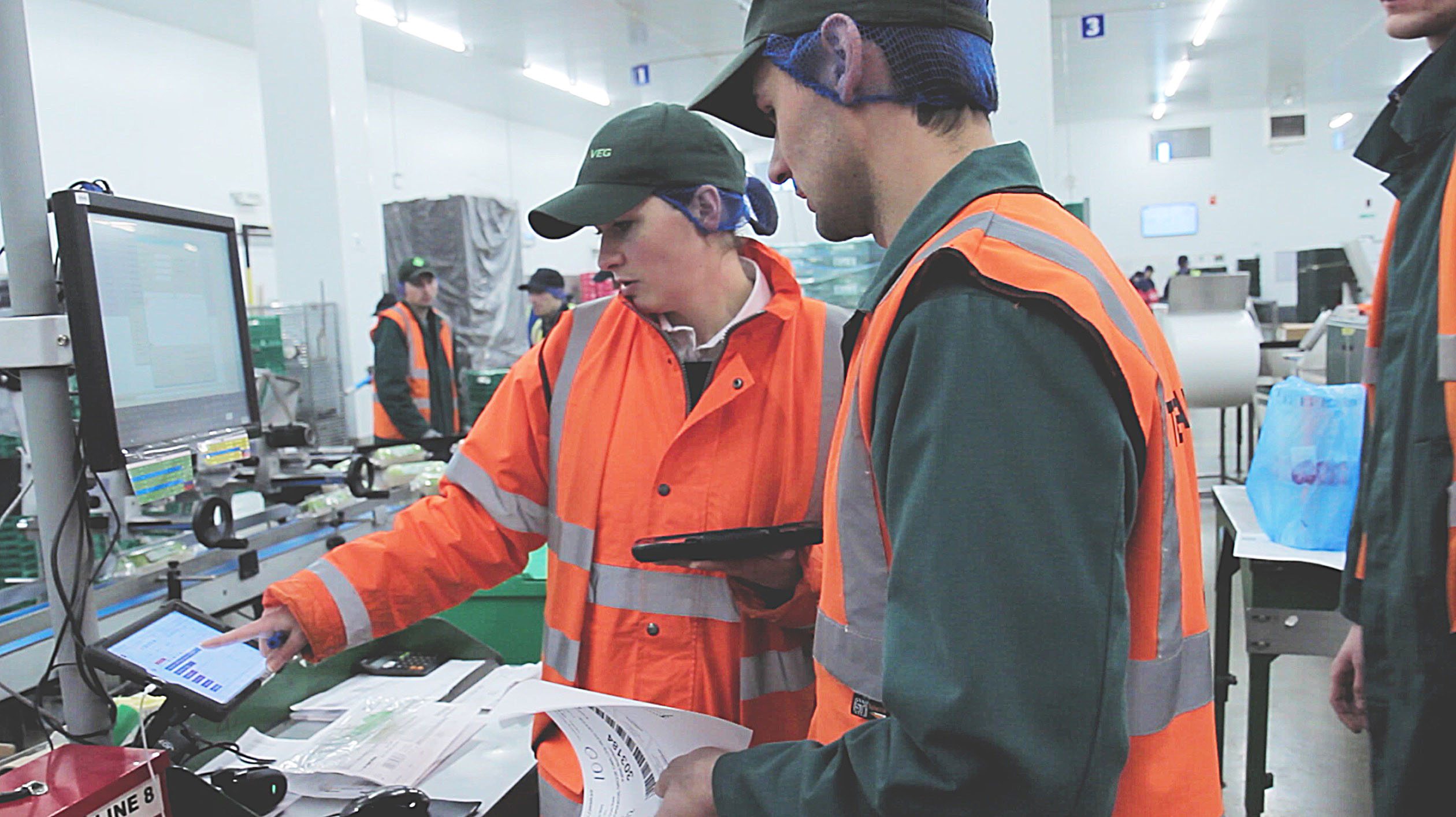 Two food processing supervisors wearing orange high-visibility jackets and hairnets review inventory data on a touchscreen terminal along a packaging line.