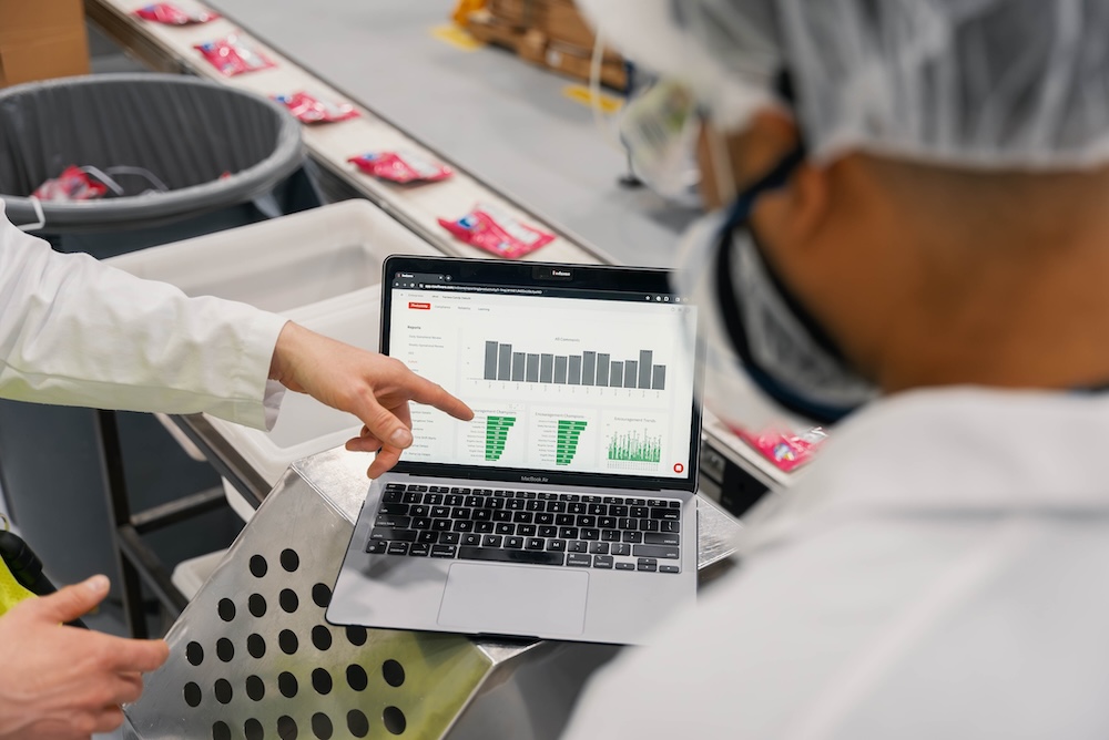 A manufacturing supervisor points to real-time analytics and performance charts on a laptop screen while reviewing production data with a lab-coated worker next to a packaging conveyor belt.