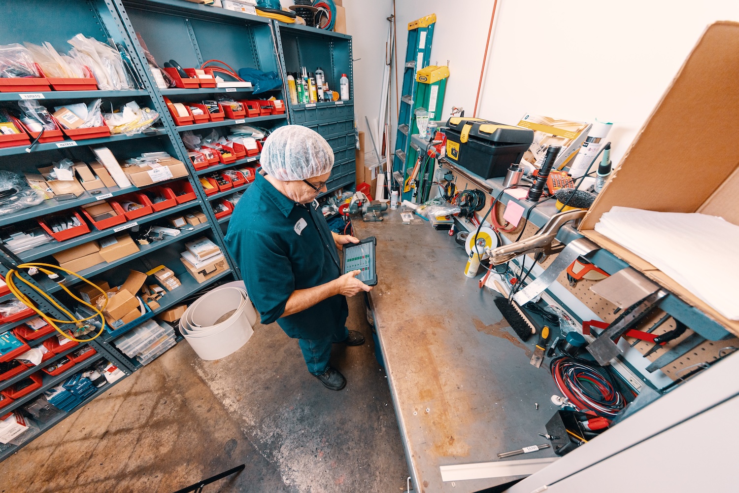 A maintenance technician wearing a hairnet reviews spare parts inventory on a digital tablet inside a organized facility toolroom with stocked shelving units.