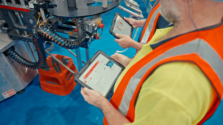 Two factory technicians in high-visibility safety vests use rugged digital tablets to monitor equipment status next to industrial production machinery.