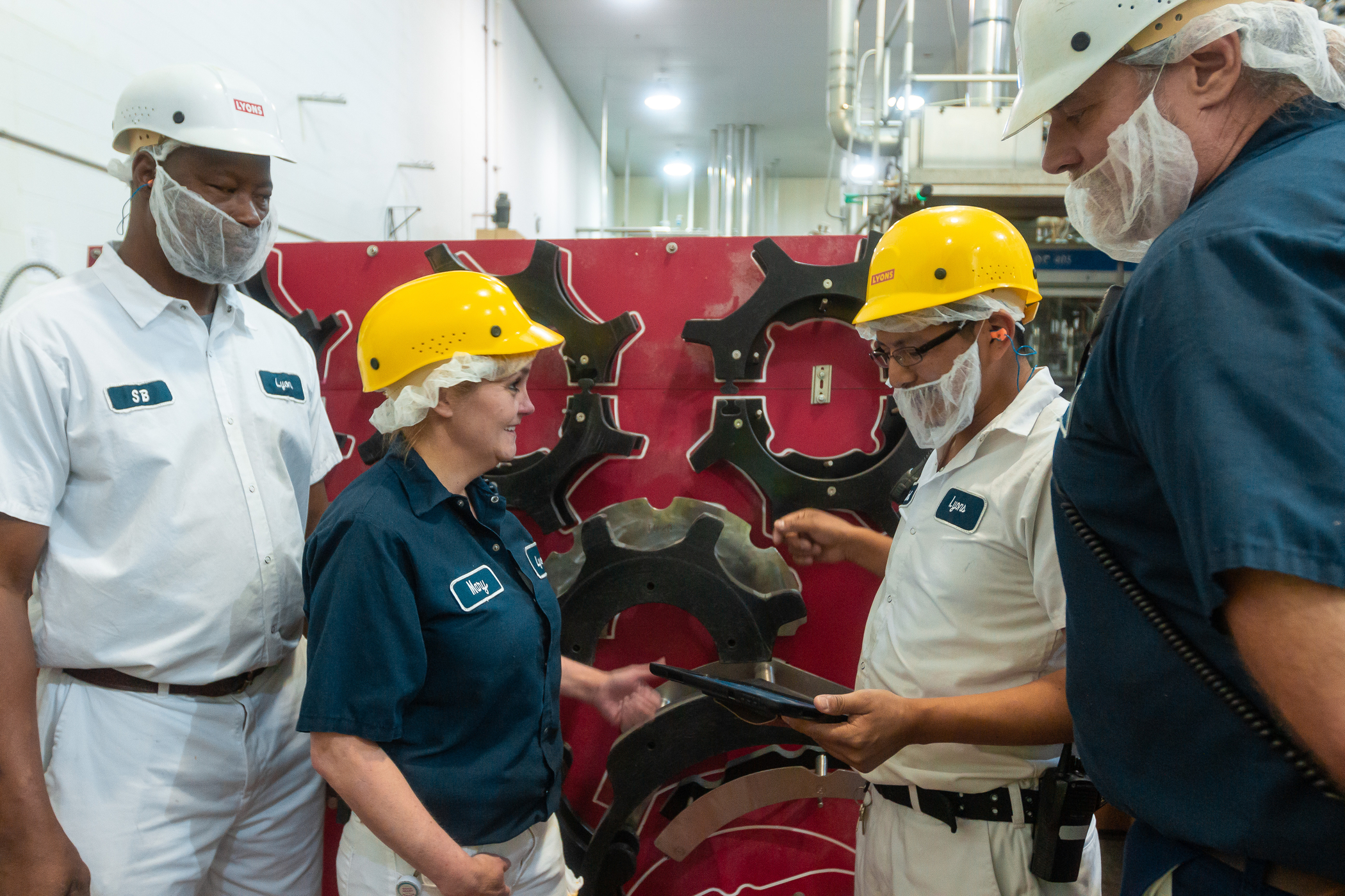 A team of maintenance workers wearing hard hats, hairnets, and beard nets discuss machinery parts while referencing a tablet near a shadow board stocked with large gears.