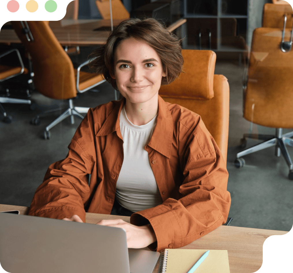Smiling woman with short brown hair wearing a rust-colored jacket sitting at a desk with a laptop in a modern office.