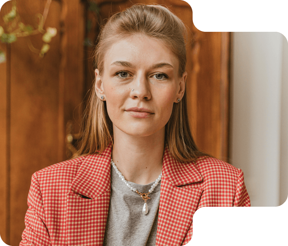 Young woman with blonde hair in a red and white checkered blazer and pearl necklace looking straight at the camera.