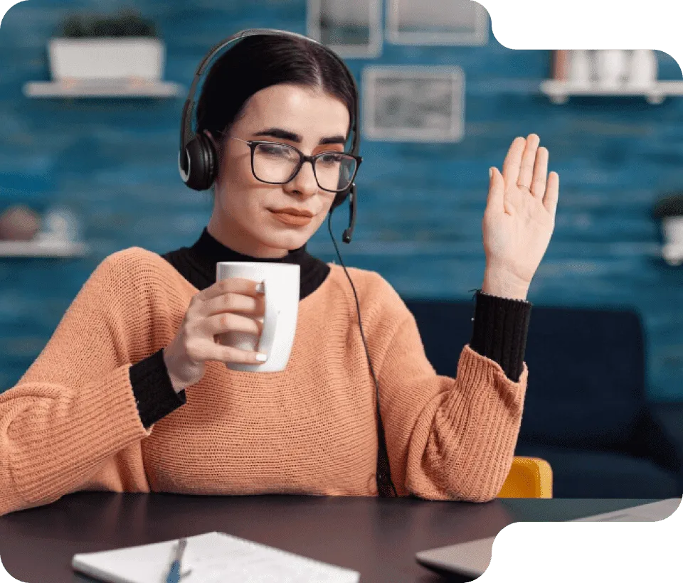 Woman wearing glasses and headset, holding a white mug and raising her hand while sitting at a desk with a laptop and notebook.