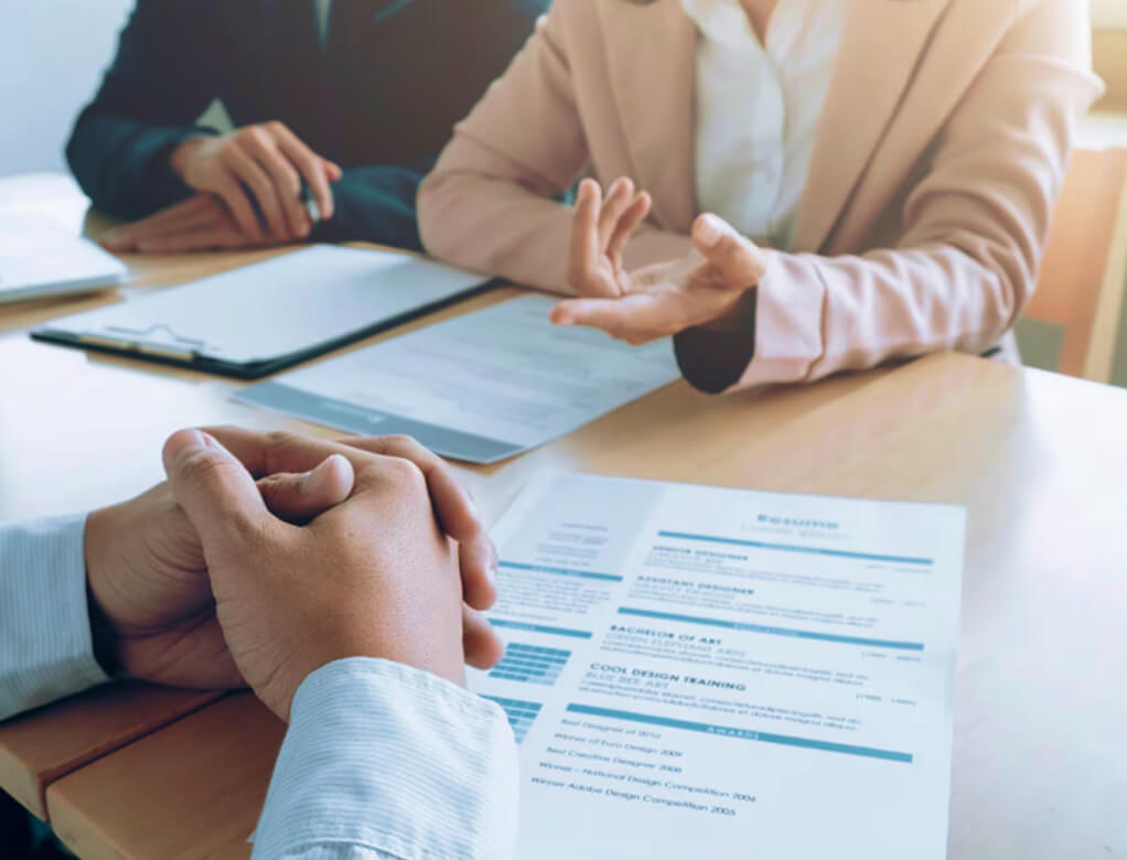 Smiling woman with short brown hair wearing a rust-colored jacket sitting at a desk with a laptop in a modern office.