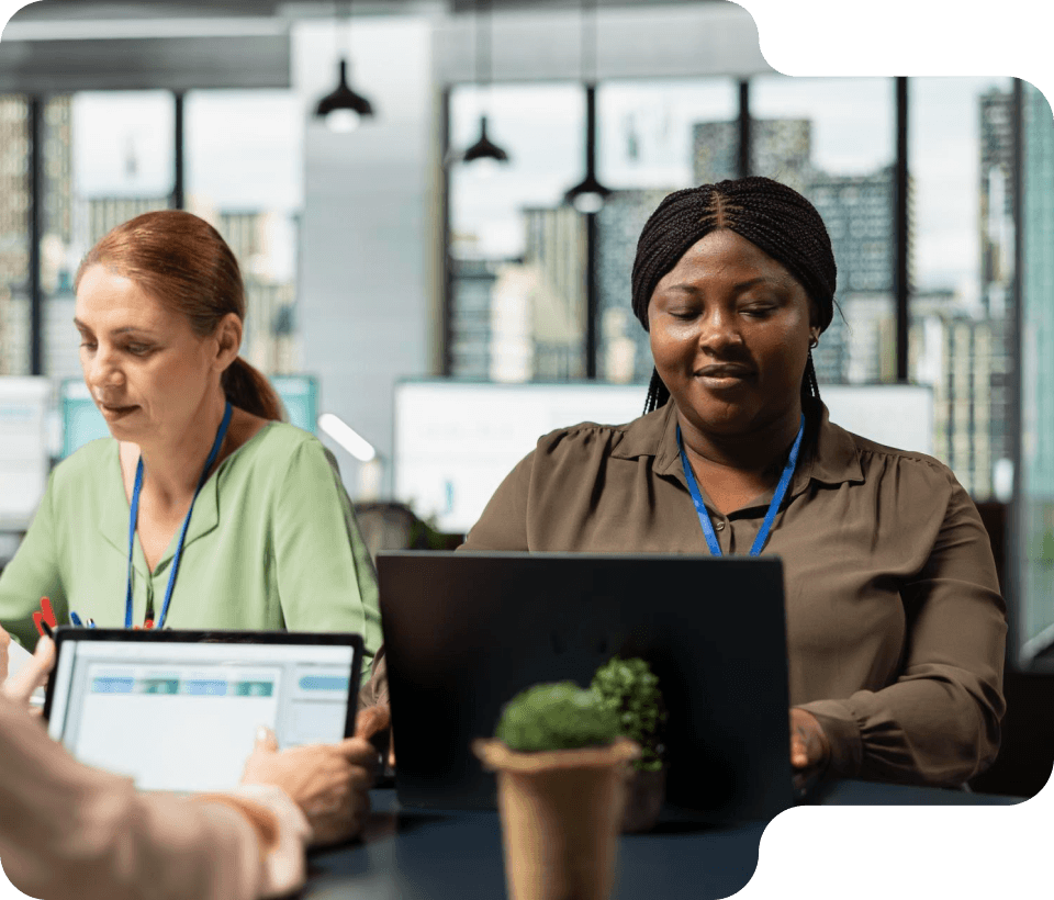 Two women working on laptops in a modern office space with city views in the background.