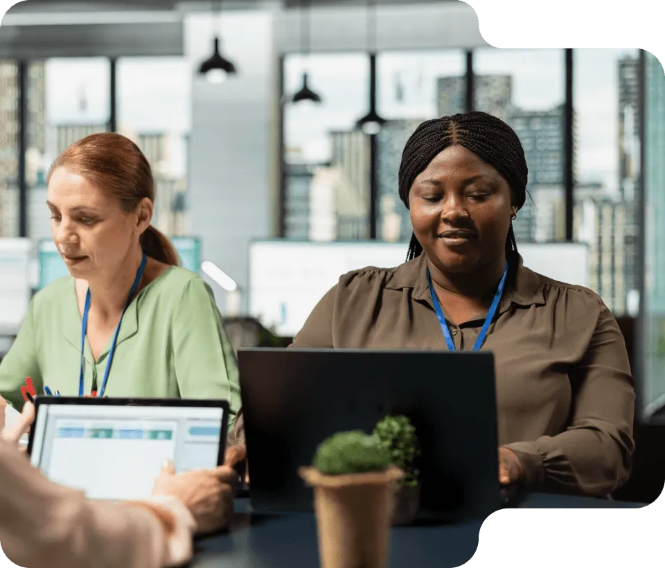 Two women working on laptops in a modern office space with city views in the background.