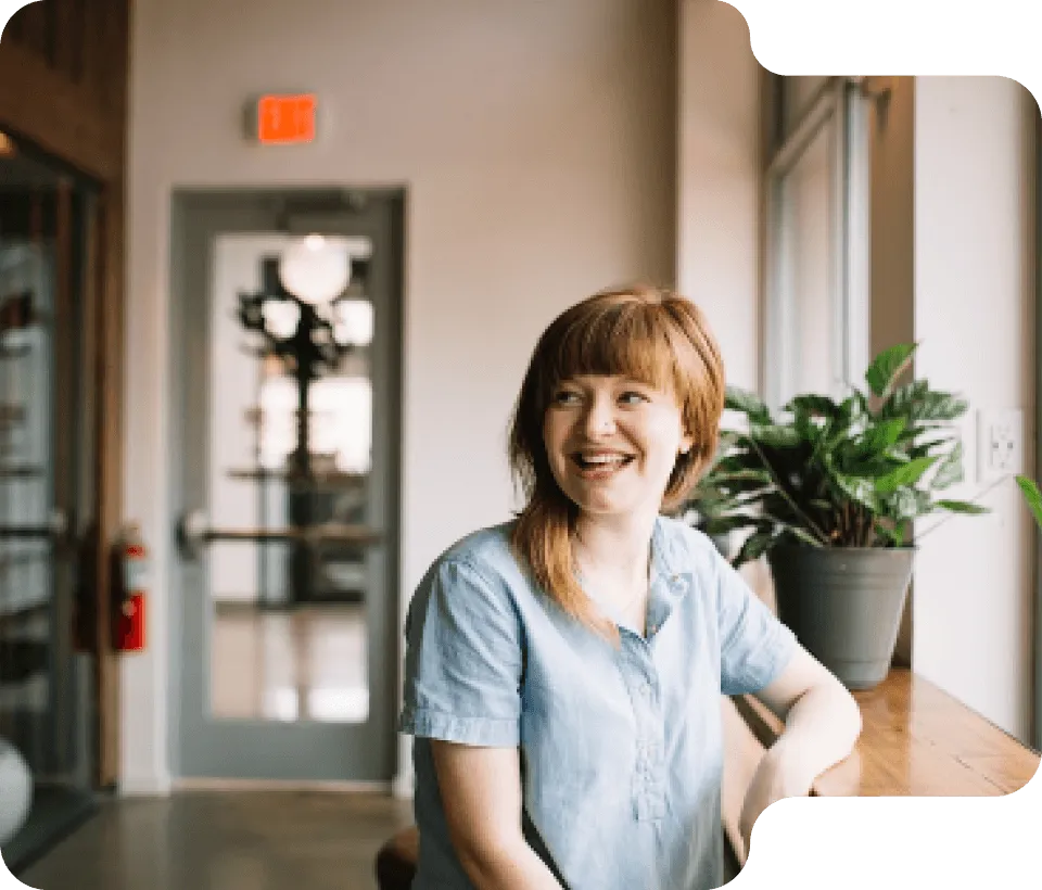 Smiling woman with red hair and blue shirt sitting by a window with a potted green plant on the wooden ledge, in a bright modern office space.