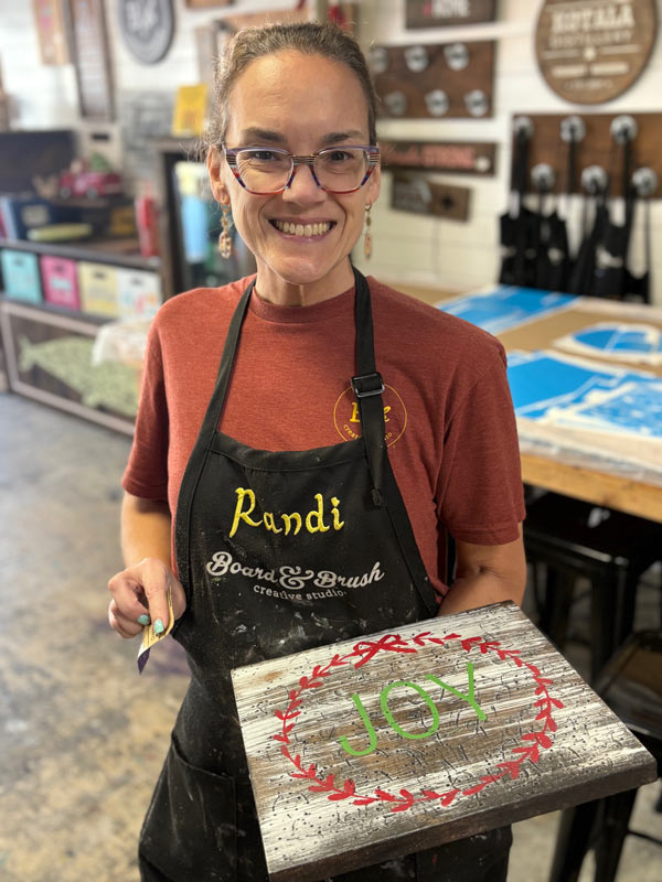 Woman holding a painted wooden sign that reads joy in a creative workshop
