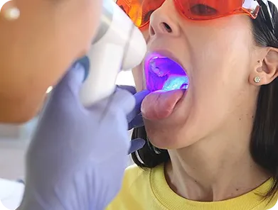 Patient wearing protective glasses has mouth open while dental professional uses a blue light device inside.