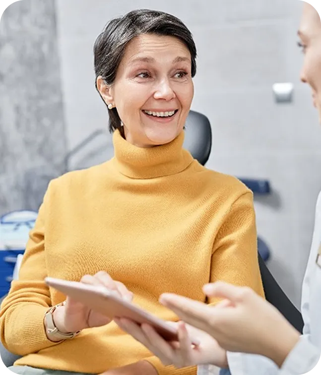 Smiling woman in a mustard yellow turtleneck discussing with a healthcare professional who is showing her a tablet.