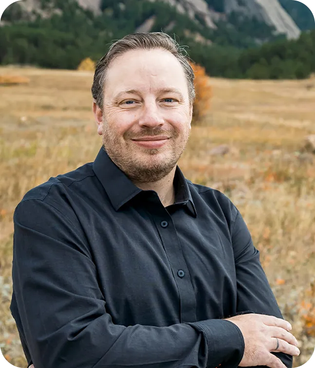 Smiling Dr Jeramiah Paylor in a black button-up shirt standing outdoors with arms crossed in a field with trees and mountains in the background.