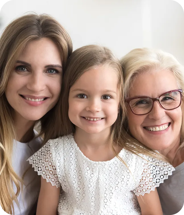 Three generations of women smiling closely together: a young girl in the center, flanked by a younger woman on the left and an older woman with glasses on the right.