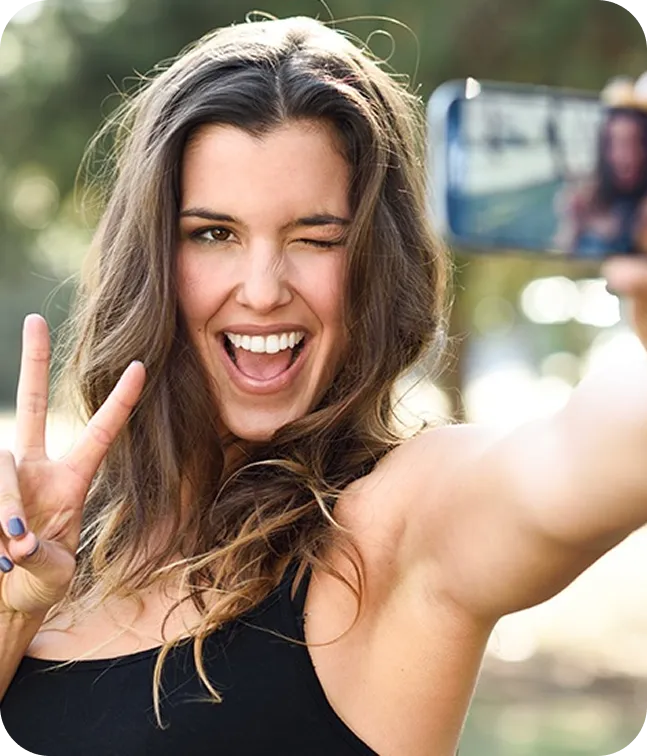 Smiling woman with long brown hair winking and making a peace sign while taking a selfie outdoors.