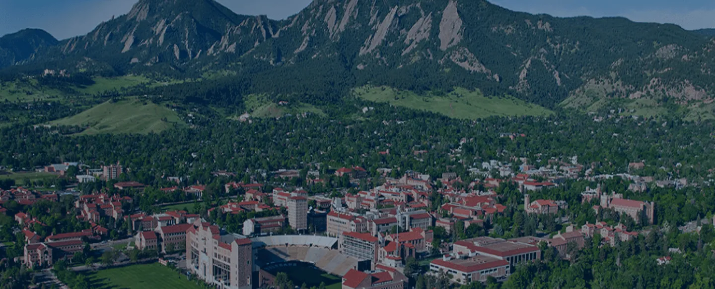 Aerial view of a university campus with red-roofed buildings surrounded by trees, set against a backdrop of forested mountains with rocky cliffs.