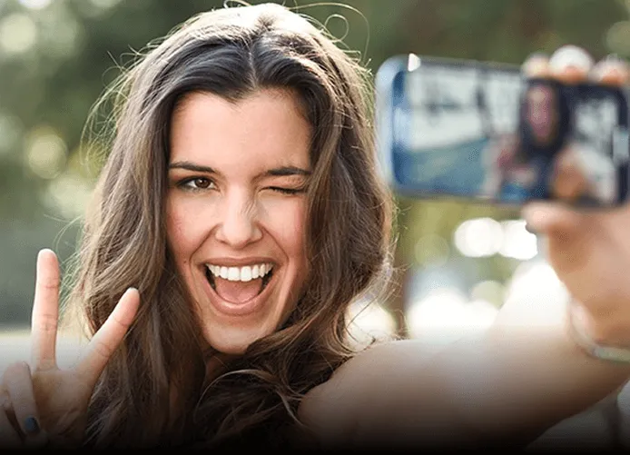 Young woman with long brown hair winking and smiling while taking a selfie outdoors, holding up a peace sign.