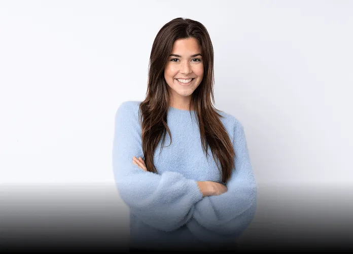 Young woman with long brown hair winking and smiling while taking a selfie outdoors, holding up a peace sign.