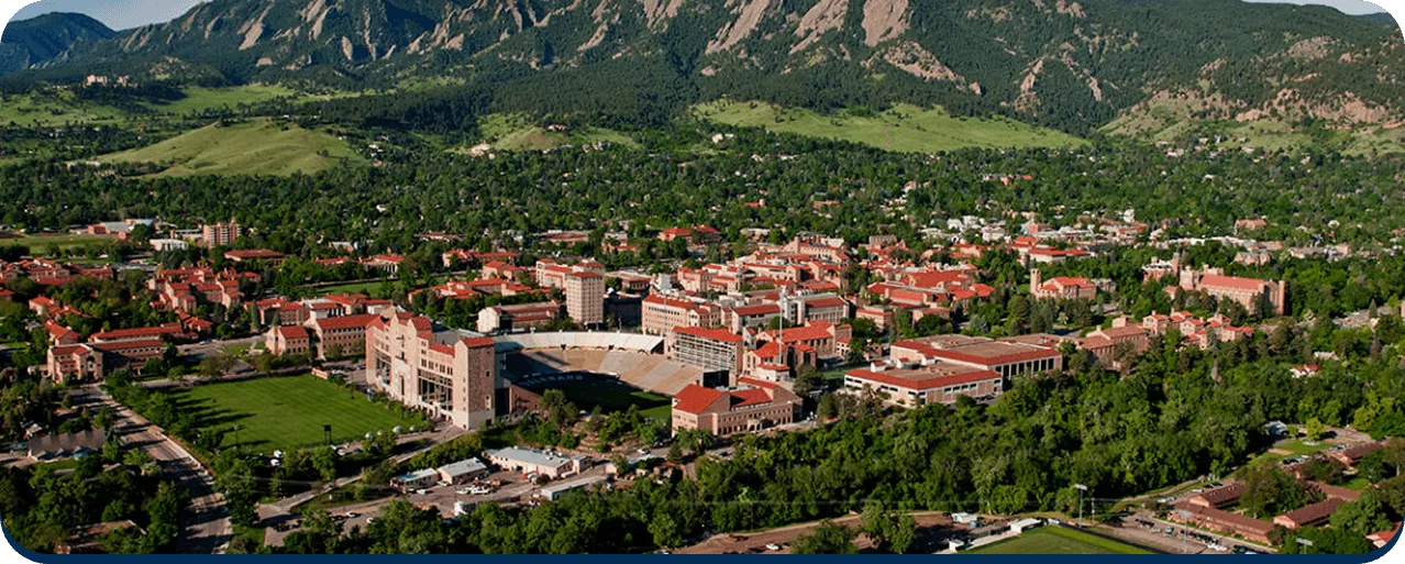 Aerial view of a university campus with red-roofed buildings, a large stadium, surrounded by trees and mountains in the background.