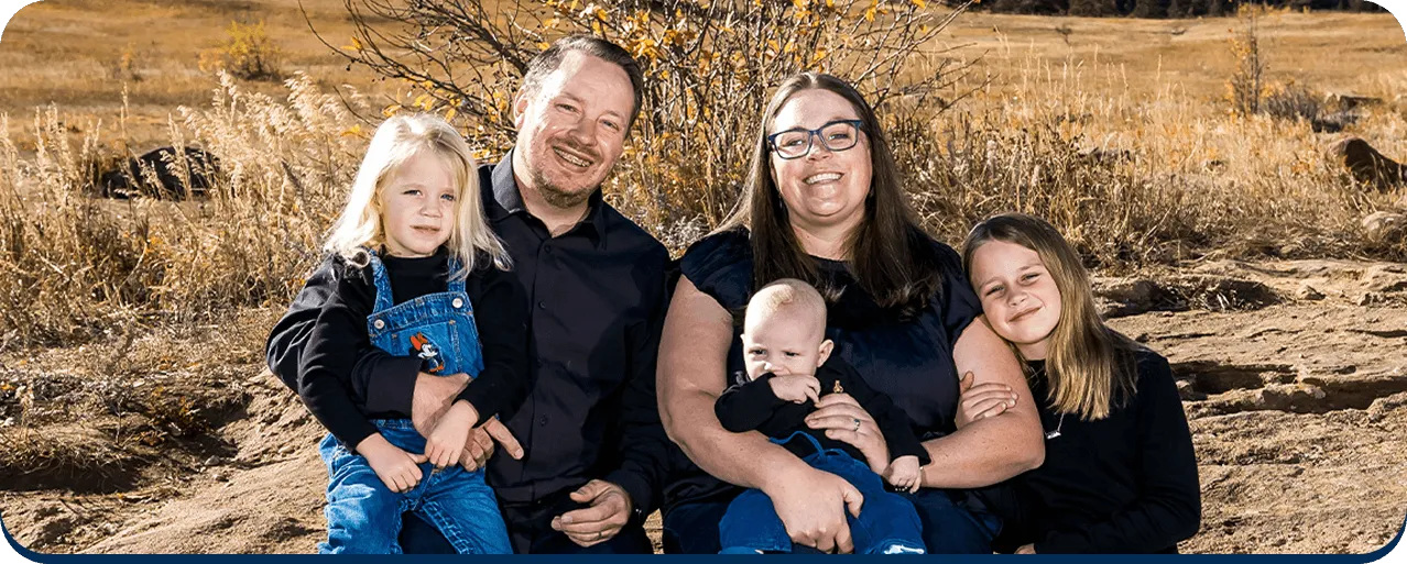 Smiling family of five sitting outdoors on rocky terrain with dry grass in the background.