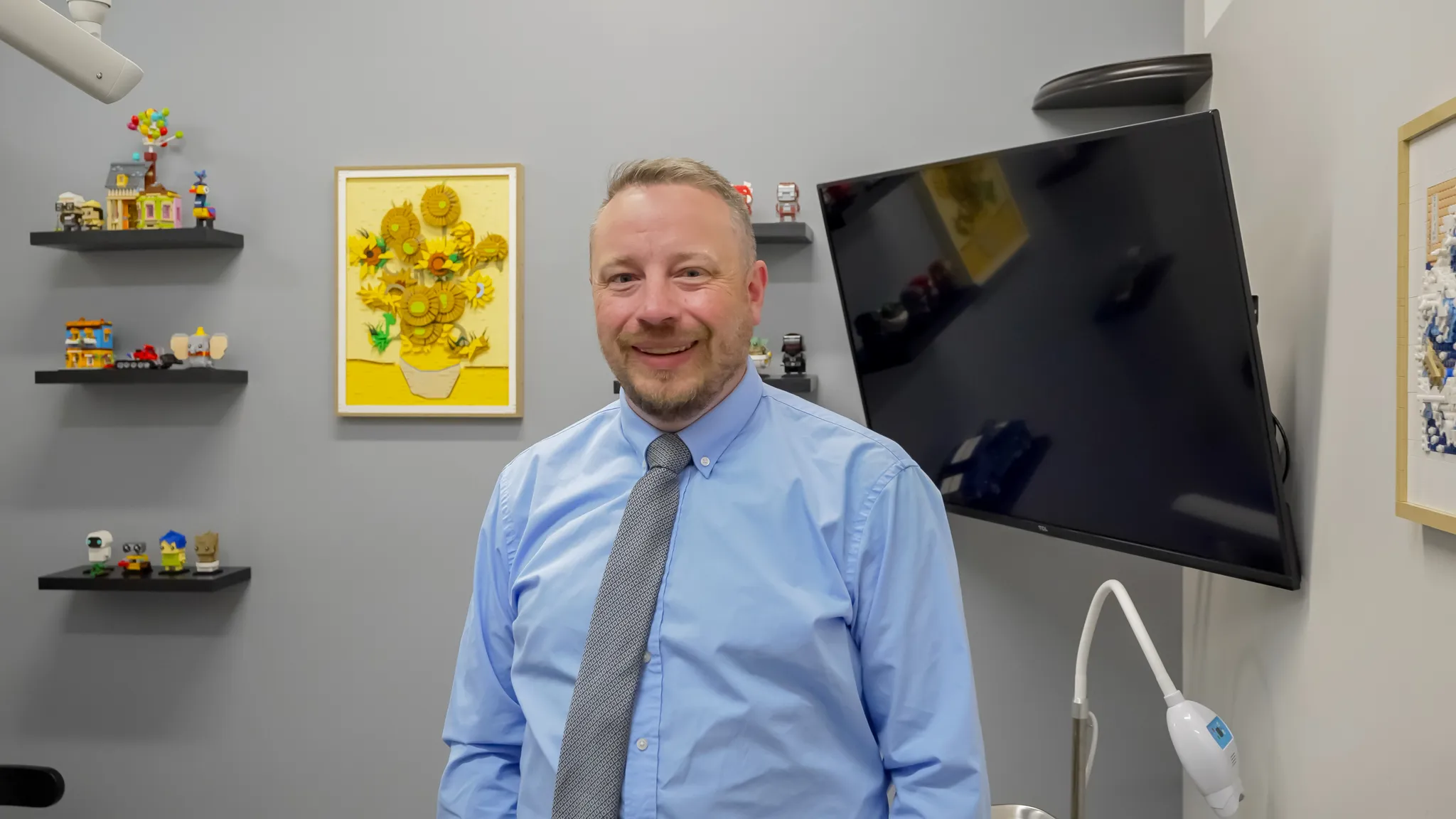 Modern dental examination room with a black and white dental chair, dental equipment, a large mirror on gray wall, and a mounted TV screen.