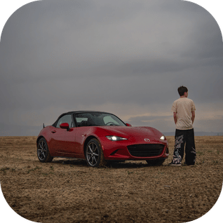 Red convertible sports car parked on a barren field with a person standing nearby under cloudy sky.