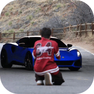 Person kneeling on road facing a blue sports car on a curved mountain road with wooden guardrails.