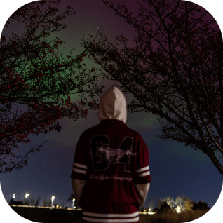 Person in hoodie and jersey standing outdoors at night under colorful northern lights.